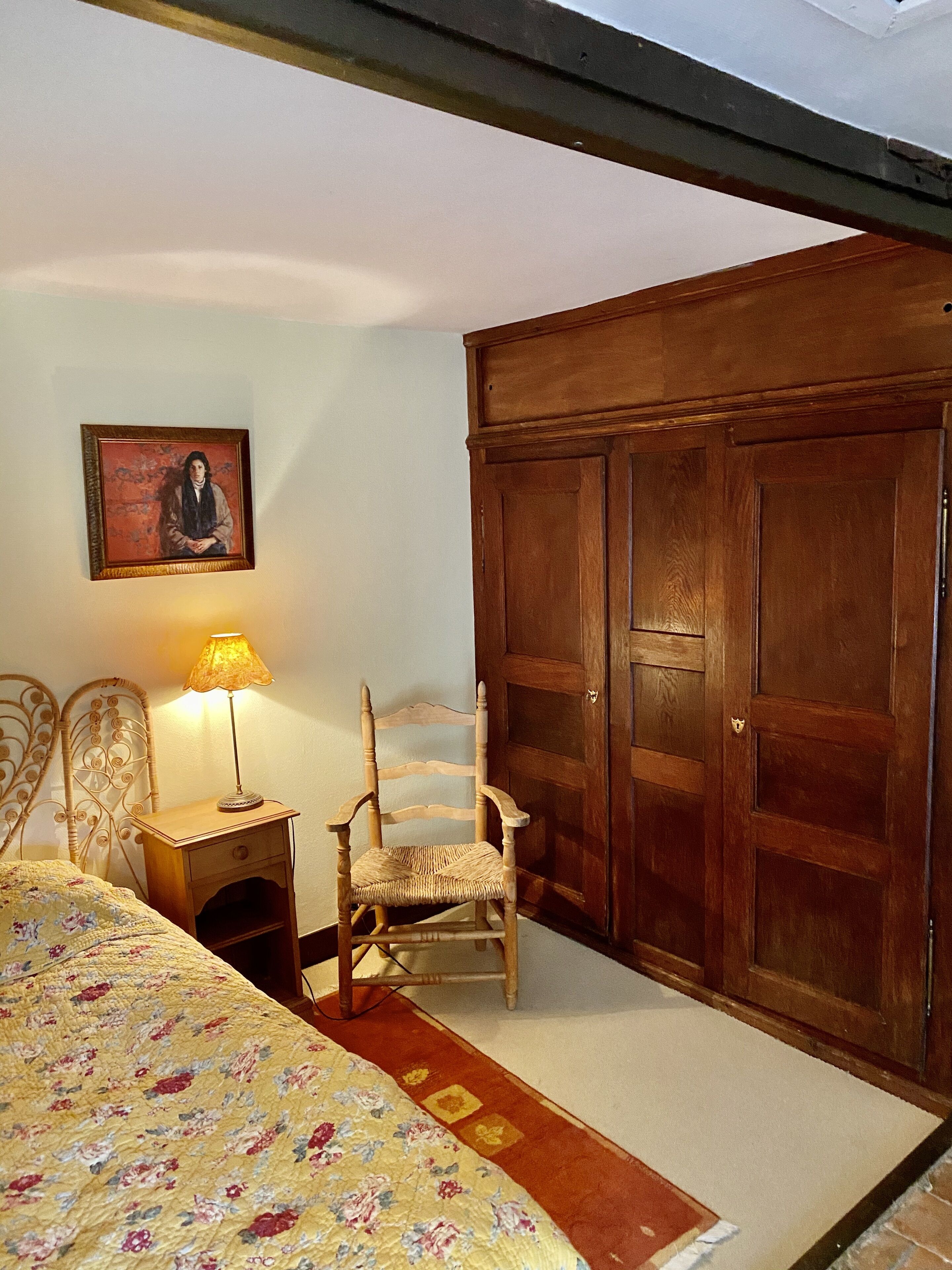 A bedroom with a bed, a wooden chair, a nightstand with a lamp, a wardrobe, and a framed picture on the wall.