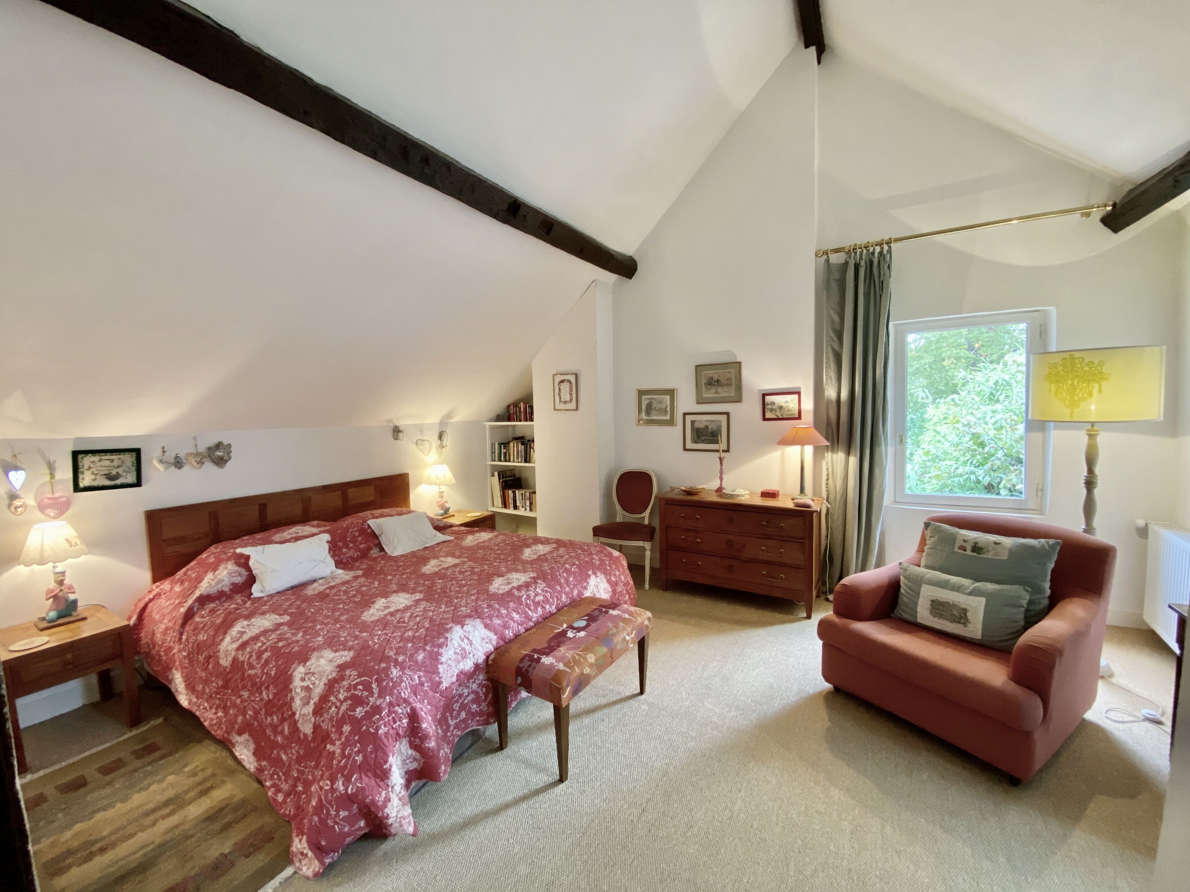 Une chambre à coucher avec un grand lit, un banc en bois, un fauteuil rouge, un bureau, une bibliothèque et une fenêtre avec des rideaux.