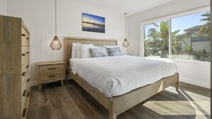 Een slaapkamer met een houten bed, nachtkastje, kledingkast en een groot raam met uitzicht op palmbomen.