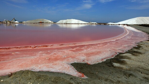 Een zoutvlakte met roze zoutwaterpoelen en witte zoutstapels onder een heldere lucht.