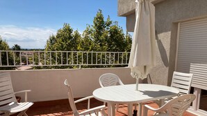 Eine Terrasse mit einem runden Tisch und vier weißen Stühlen, einem weißen Sonnenschirm und Blick auf Bäume und Gebäude im Hintergrund.