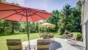 A wooden deck with wicker chairs and a table, surrounded by a lush garden and trees. - Heron Pointe... where nature and luxury combine for an unforgettable experience! (White Lake)
