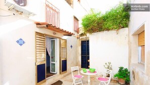 A narrow courtyard with a white table and chairs, a potted plant, and a window with shutters.