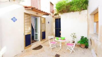 Une petite cour étroite avec une table et des chaises blanches, un pot de fleurs et une fenêtre avec des volets.