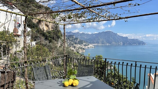 A terrace with a table and chairs, overlooking a coastal view with mountains and a body of water.