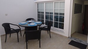 Een klein terras met een ronde tafel voor twee personen, omringd door stoelen, en een groot raam met zonwering.