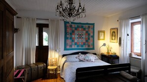 Un dormitorio con cama, candelabro, colcha, silla, maleta y ventana con cortinas.