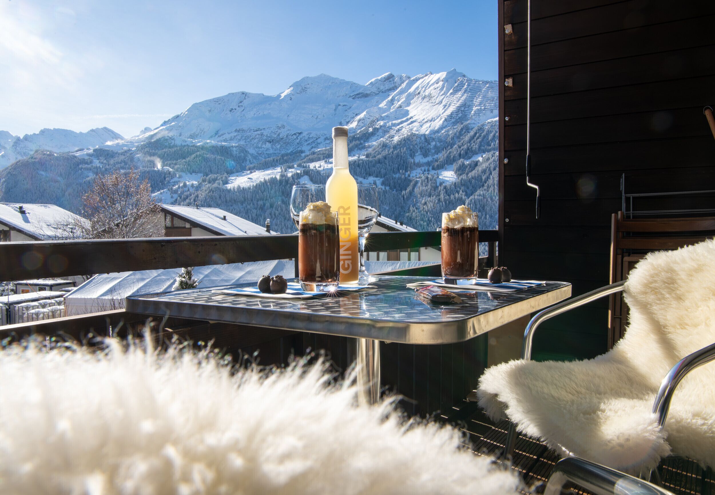 Uma mesa com uma garrafa de cerveja de gengibre, dois copos e uma tigela, ao fundo de uma paisagem montanhosa coberta de neve.