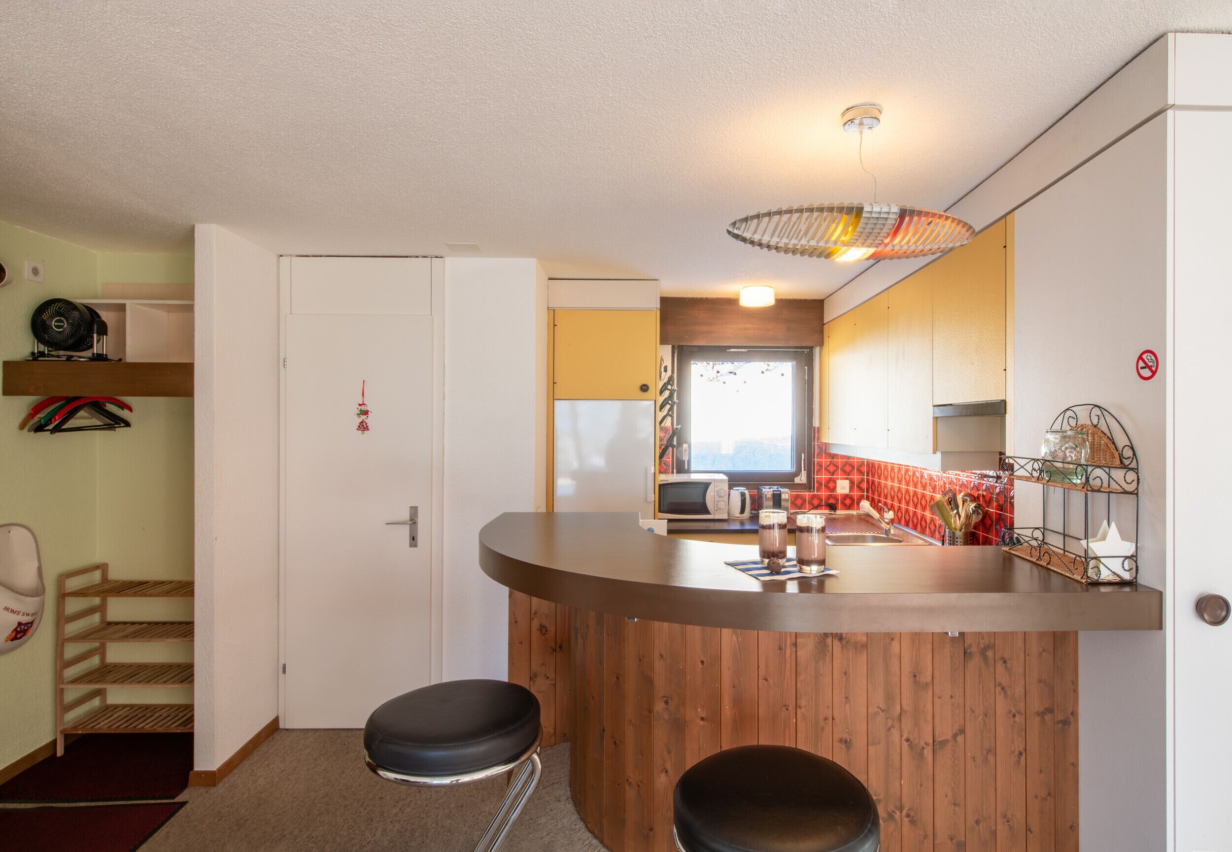 Eine kompakte Küche mit einer Holzbar, einer Mikrowelle und einem Spülbecken. Zwei Hocker und ein kleines Regal mit Küchenutensilien sind vorhanden.