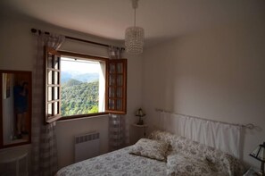 Quarto com uma janela grande, cama com roupa de cama estampada, mesa de cabeceira e luminária pendente.