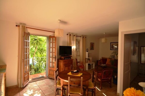 Uma sala de jantar com uma mesa redonda e cadeiras, uma televisão sobre um suporte de madeira, e uma varanda com vista para o verde.