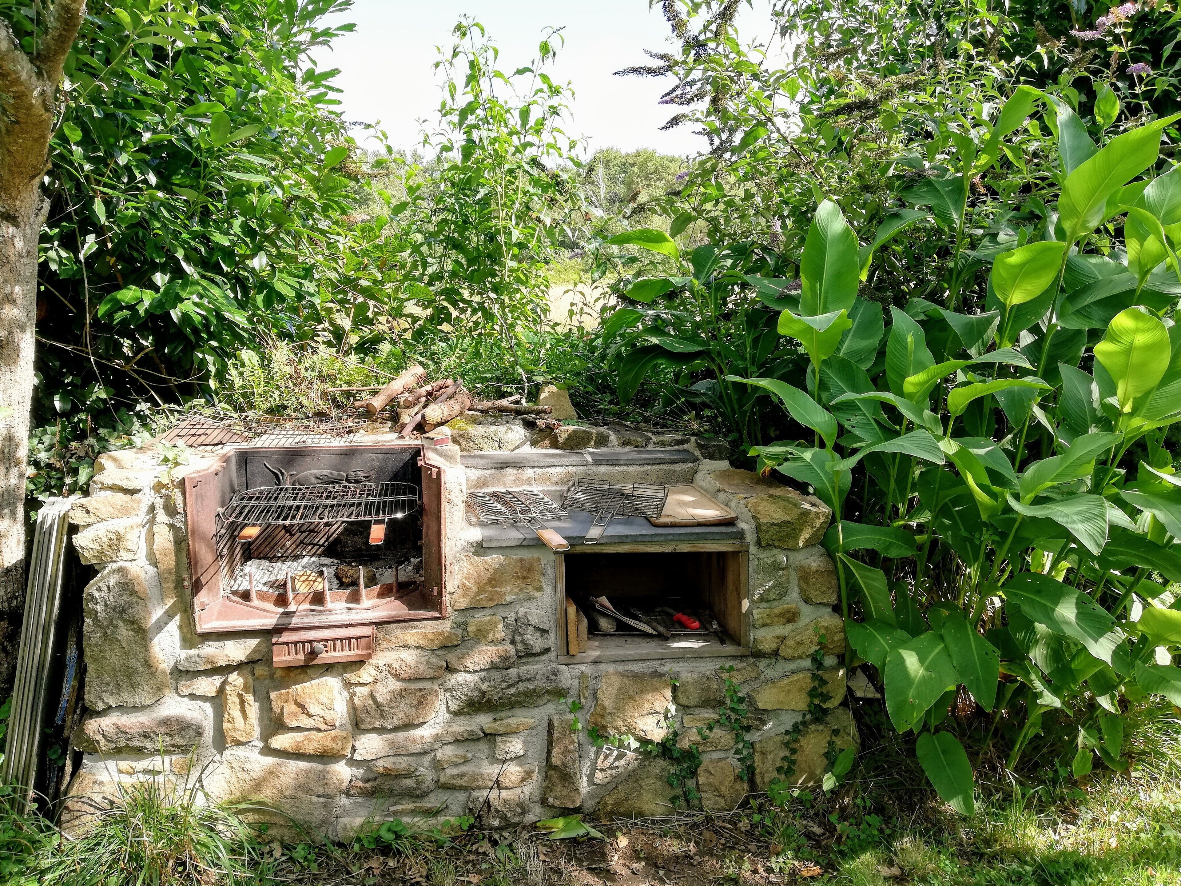 Un barbecue extérieur en pierre, doté de plusieurs surfaces de cuisson et d’une cheminée, entouré d’une végétation luxuriante.