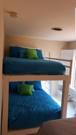 Un lit superposé avec une literie bleue et des oreillers verts, dans une pièce dotée d’une fenêtre et d’une armoire blanche.