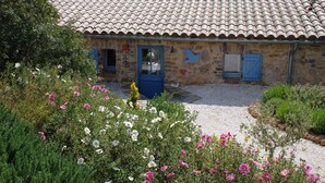 Un bâtiment en pierre avec un toit en tuiles, une porte bleue et un jardin où poussent différentes sortes de fleurs.