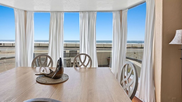 A dining area with a wooden table and chairs, large windows with white curtains, and a view of the beach and ocean.