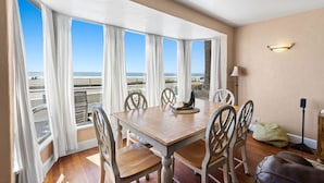 A dining area with a wooden table and chairs, large windows with white curtains, a lamp, and a view of the beach.