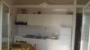 Een kleine keuken met witte kastjes, een spoelbak en een koelkast. Op de voorgrond staat een eettafel met stoelen.