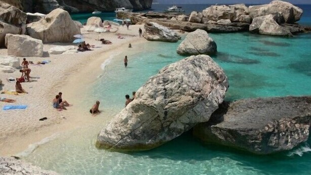 Een strand met grote rotsen, helder turquoise water en mensen die zonnen.