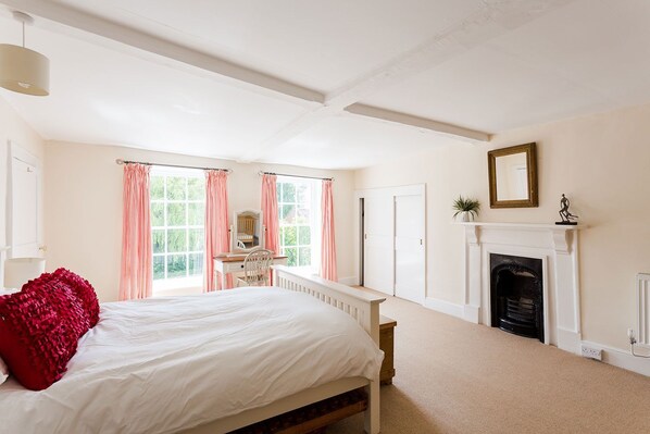 A bedroom with a large bed, a desk with a chair, a fireplace, and a window with curtains.