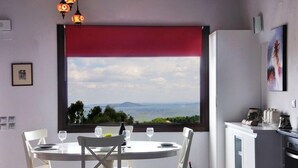 A dining area with a table set for four, a window with a red blind, and a view of the landscape. - Alonissos, Old Village (Hora) wonderful apartment with amazing  clear sea views (Alonissos)