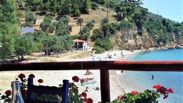 A table with a bottle and glass, overlooking a beach with people and a building in the background. - Alonissos, Old Village (Hora) wonderful apartment with amazing  clear sea views (Alonissos)
