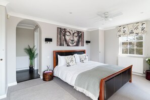 A bedroom with a bed, a ceiling fan, a window with curtains, a wall-mounted artwork, and a potted plant.