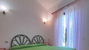 A bedroom with a green bedspread, a metal headboard, a bedside table, and a window with sheer curtains.
