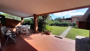 A covered patio area with a table and chairs, a grill, and a view of a well-maintained lawn and garden.