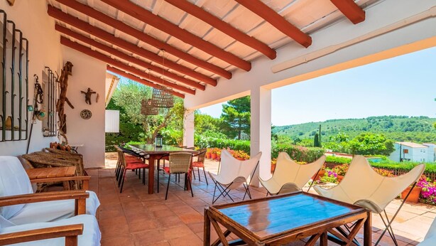 A covered patio with a table and chairs, a view of greenery and hills, and a rustic interior with wooden beams and a hanging light fixture. - Costabravaforrent Masramon (l'Escala)