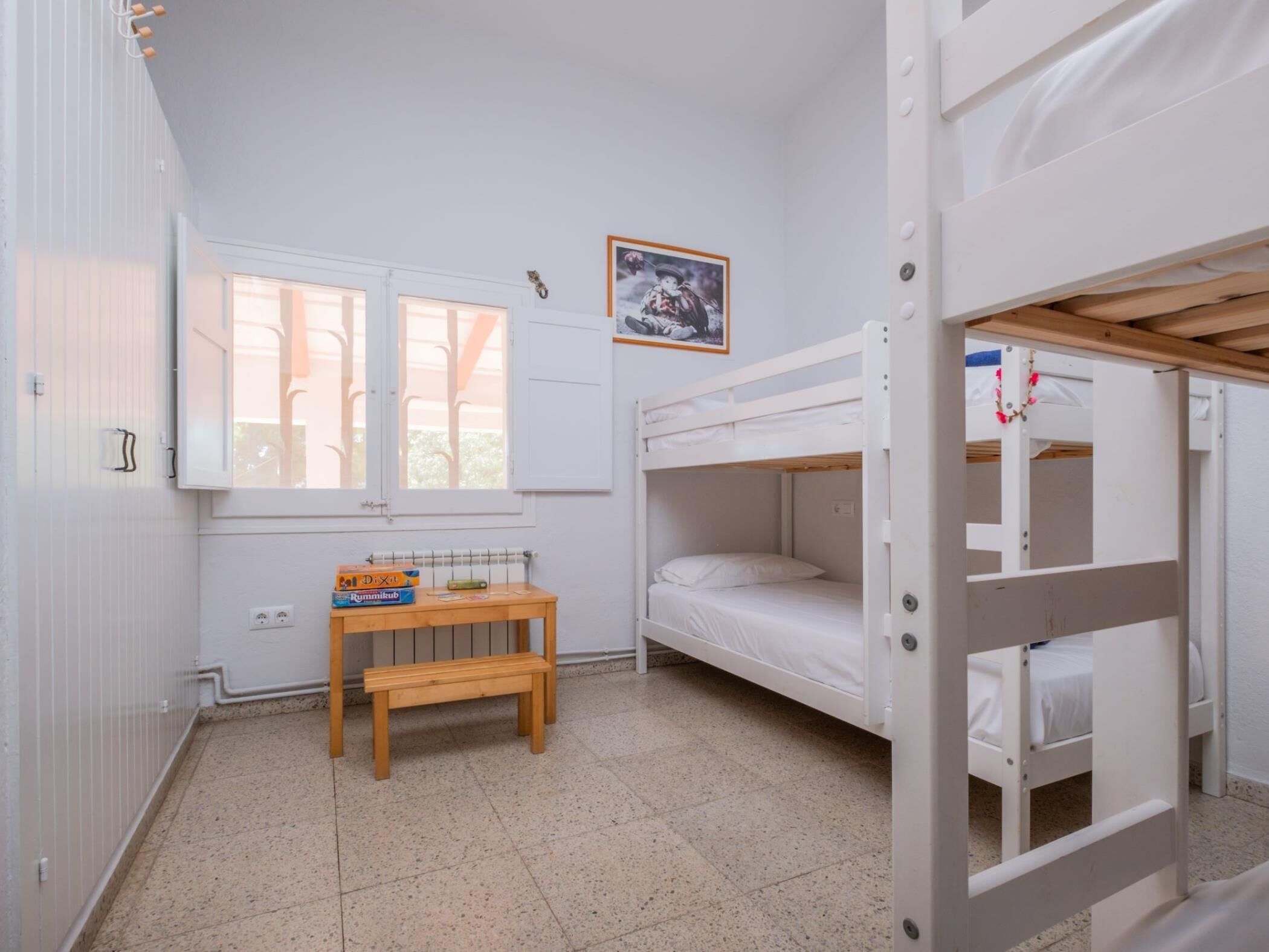 A bunk bed room with a window, a small table with books, and a framed picture on the wall.