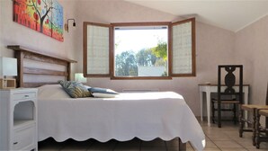 Ein Schlafzimmer mit einem Bett, einem Nachttisch, einem Sessel, einem Schreibtisch und einem Fenster mit Blick auf Bäume.