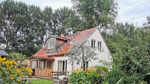 Una casa de dos pisos con techo inclinado, un porche de madera y un jardín con diversas plantas y flores.