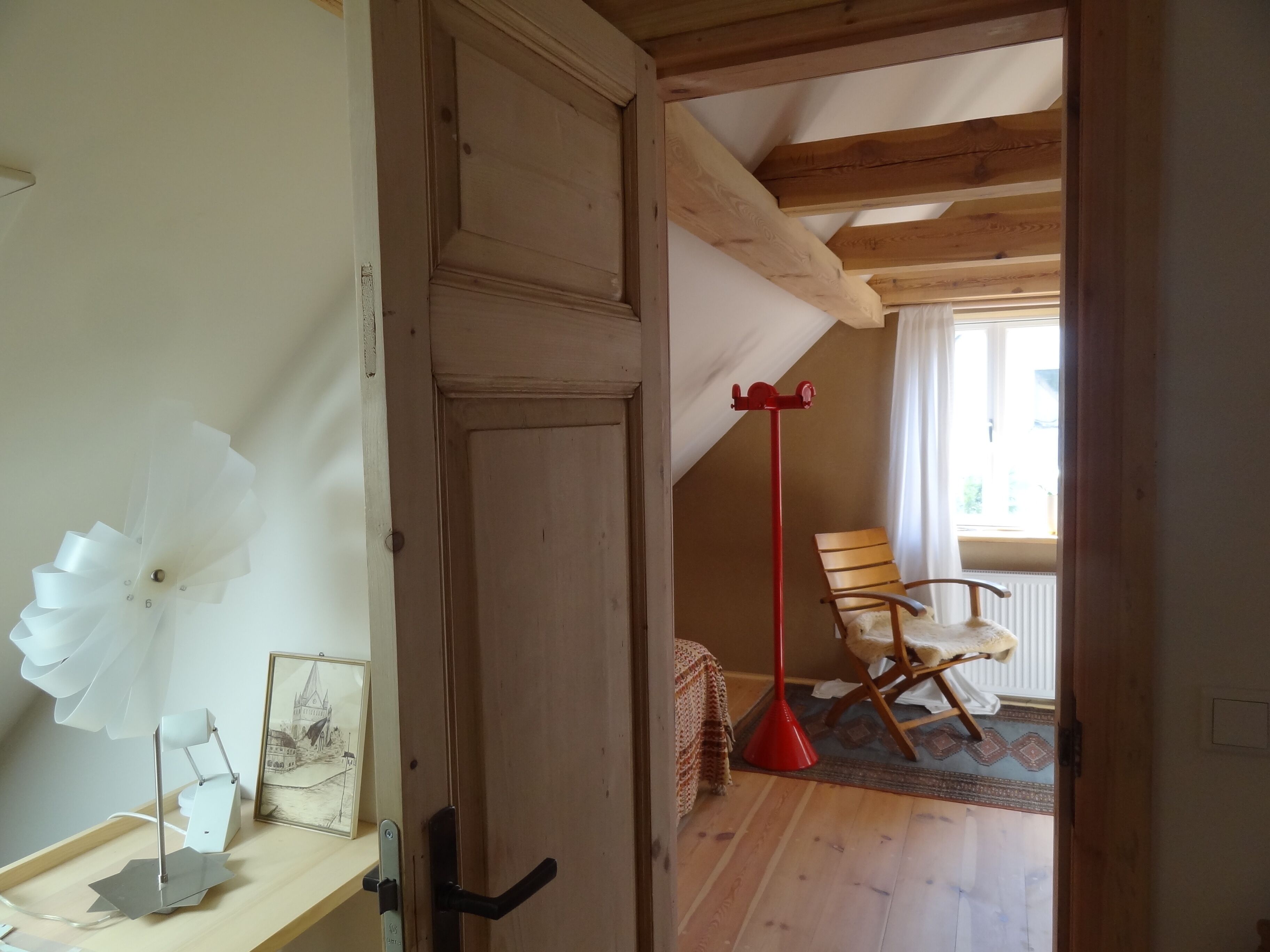 Een kamer met een houten deur, een rode vloerlamp, een houten stoel en een lijst met een foto op een plank.