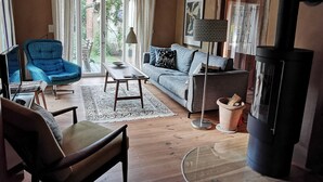 Una sala de estar acogedora con un sillón azul, un sofá, una mesa de centro, una chimenea y un ventanal con cortinas.