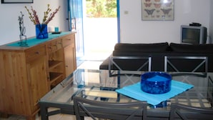 Comedor con mesa de vidrio, sillas, un florero azul con ramas, un mueble de madera, un sofá y un televisor.