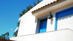 Un edificio blanco con puerta y ventana azules, una antena parabólica y una terraza de madera al frente.