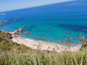 A coastal landscape with clear blue waters, rocky shoreline, and a sandy beach.