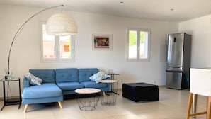 Una sala moderna con un sofá seccional azul, dos mesas redondas de centro, una lámpara colgante grande, un refrigerador y una mesa de comedor.