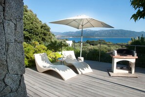 Une terrasse dotée de deux chaises longues, d’une table et d’un barbecue, offrant une vue panoramique sur la mer et les montagnes.