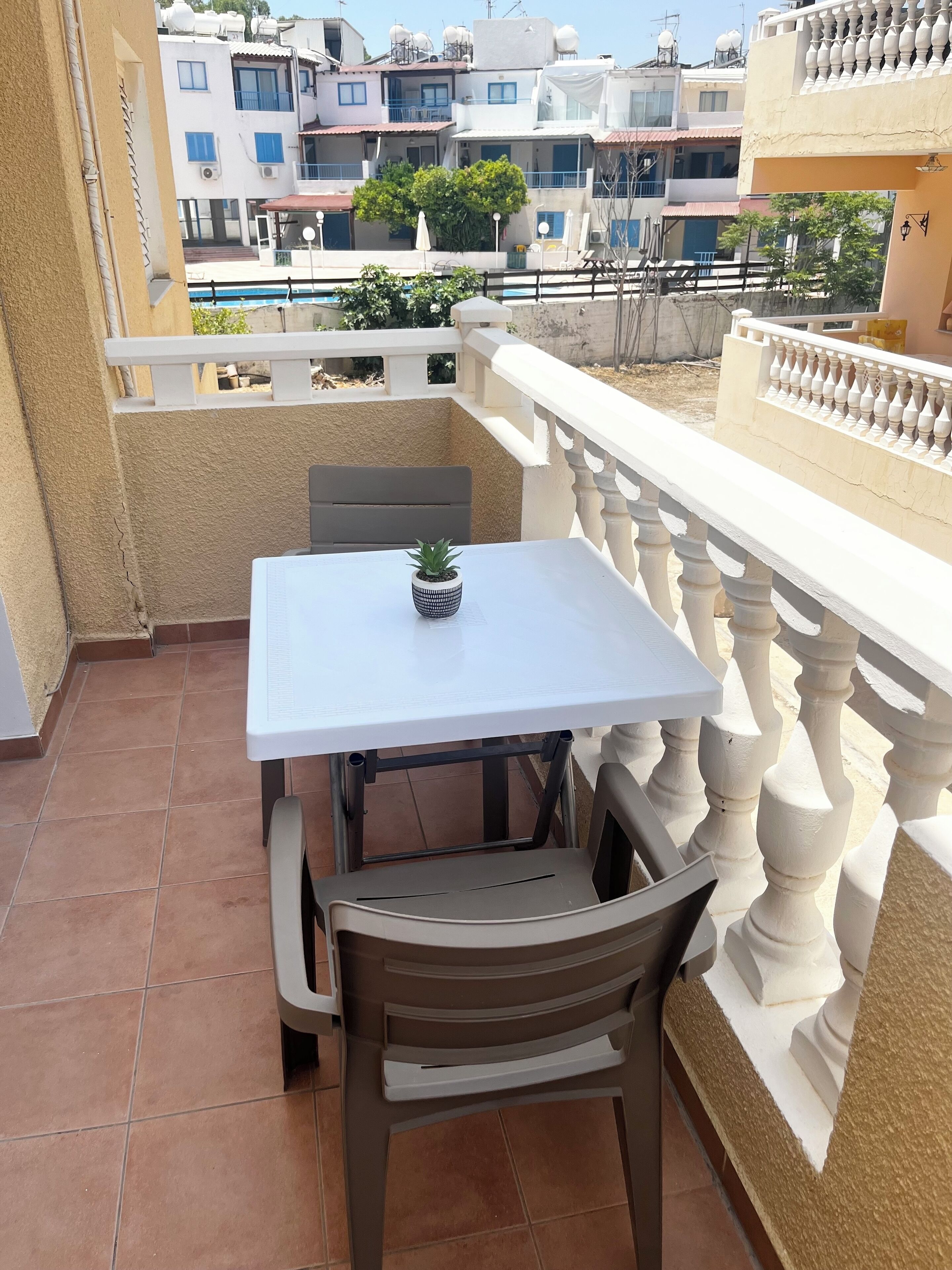 Ein Balkon mit einem weißen Tisch und einem einzelnen Stuhl, eine kleine Topfpflanze auf dem Tisch und Blick auf benachbarte Gebäude.