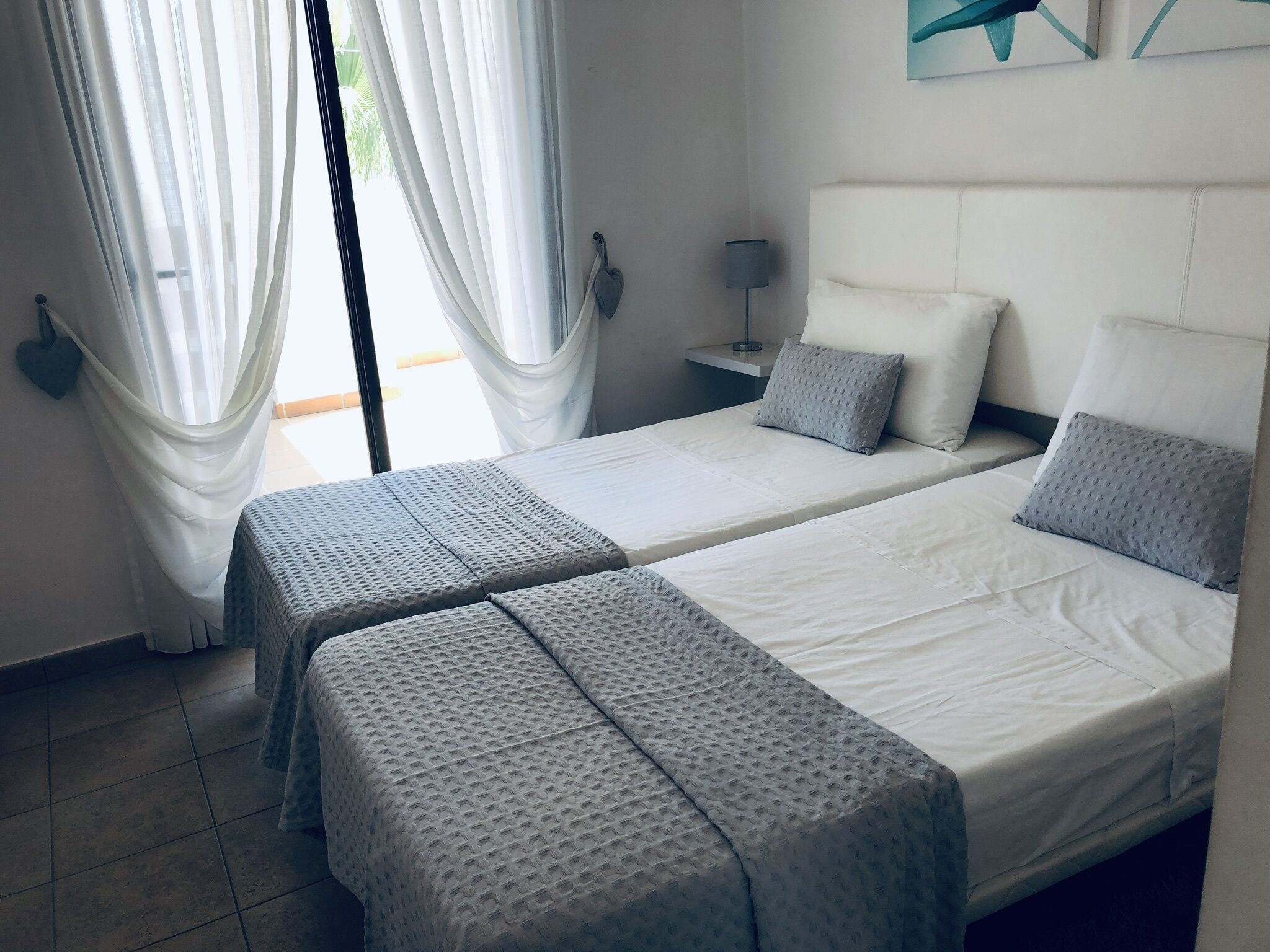 A hotel room with two beds, a nightstand, a lamp, and sheer curtains.