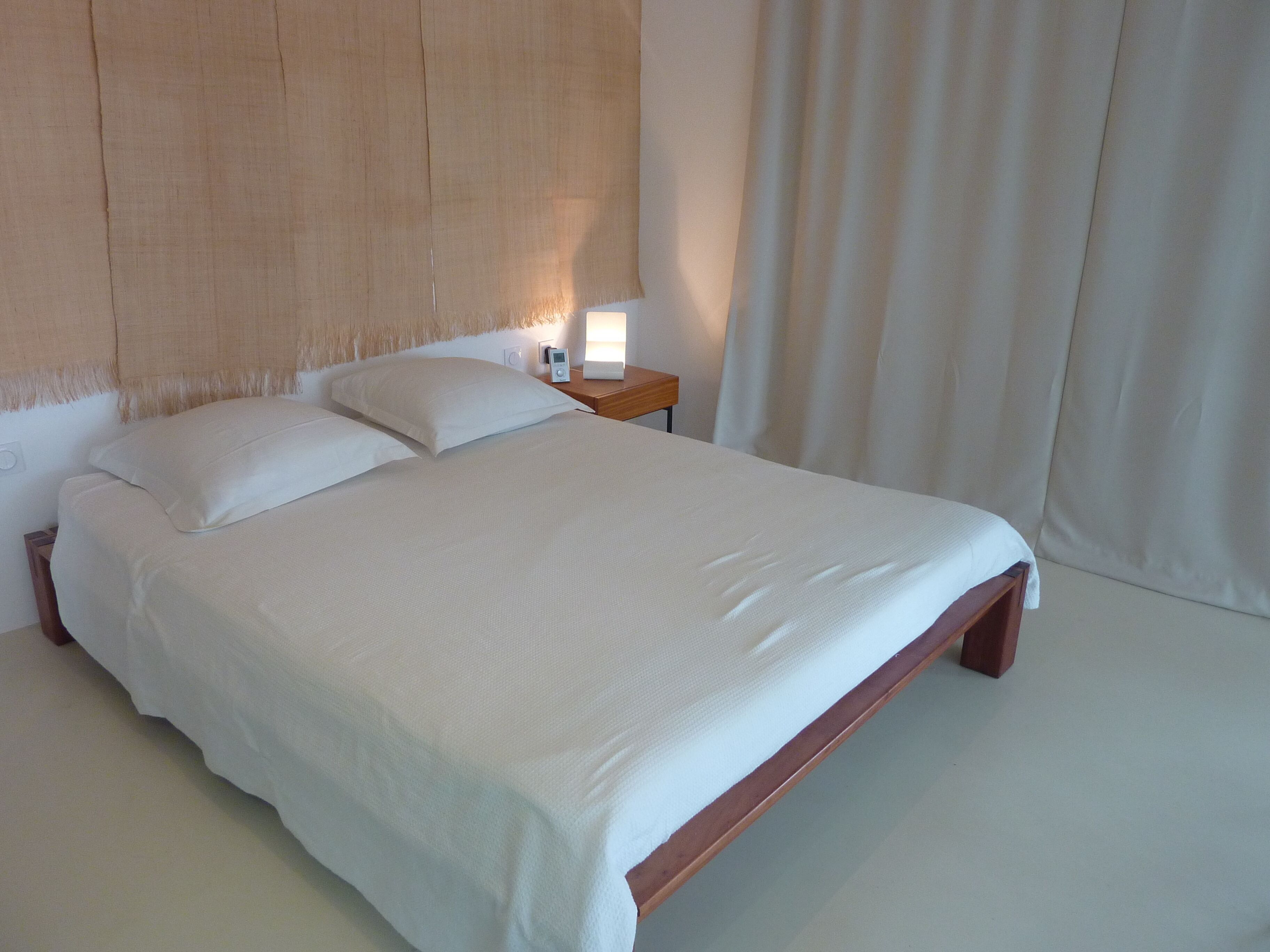 A neatly made bed with white linens, a wooden headboard, a bedside table with a lamp, and a curtain to the right.