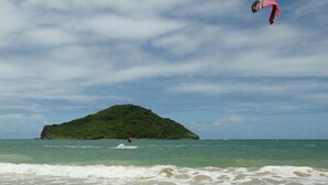 Una playa con agua cristalina, una pequeña isla y una persona practicando kitesurf.
