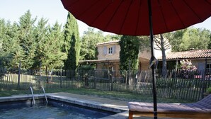 A pool area with a wooden deck, lounge chairs, and a red umbrella. - Villa in the Luberon (Apt)