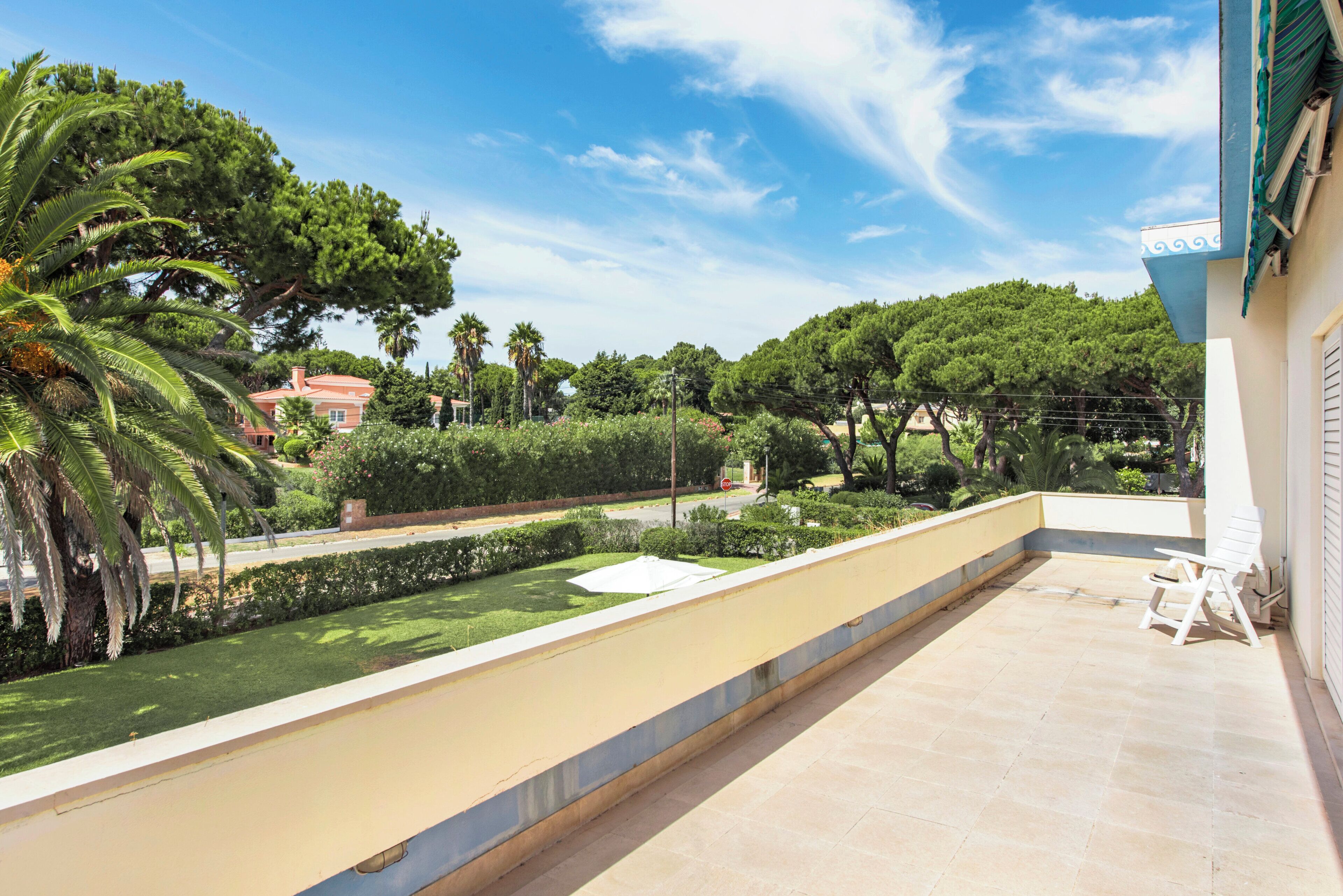 Une terrasse donnant sur un jardin et des arbres, une chaise blanche et un bâtiment surmonté d’un auvent bleu.