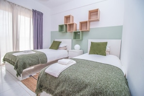 A hotel room with two beds, each with a green bedspread and white pillows, a small bedside table with a lamp, and a wall-mounted shelf above the beds.