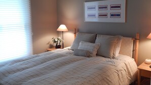 Ein ordentlich bezogenes Bett mit einer karierten Bettdecke, eine Nachttischlampe, ein gerahmtes Bild an der Wand und ein Fenster mit Jalousien.