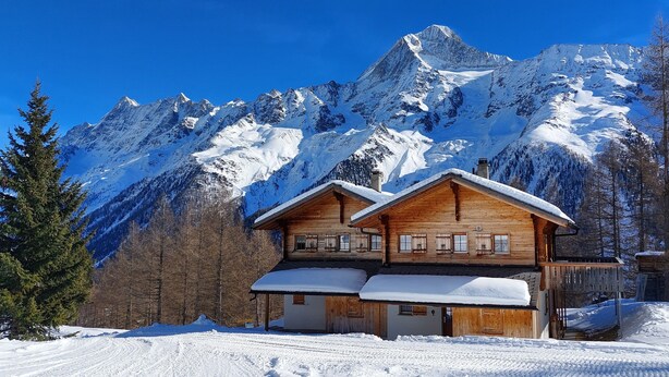 A wooden chalet with a balcony, surrounded by snow-covered mountains and trees.