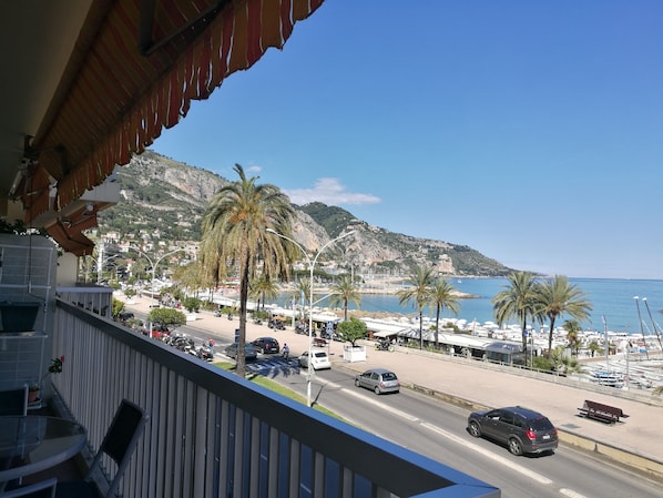 Ein Balkon mit Blick auf eine Küstenstraße, wo Autos, Palmen und das Meer sowie Berge zu sehen sind.