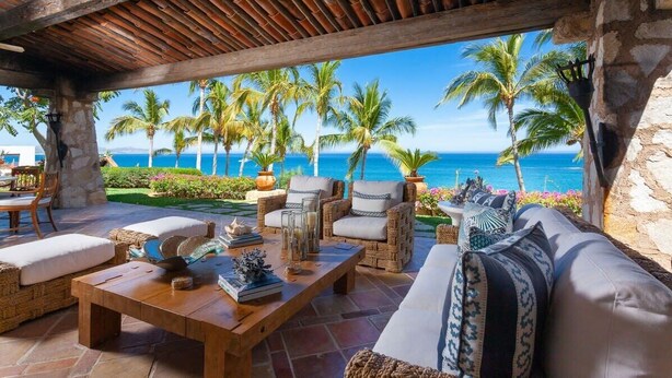 An outdoor seating area with wicker furniture, a wooden table, and a view of the ocean and palm trees. - Beachfront Paradise! Please see instructions below prior to booking! (San Jose del Cabo)